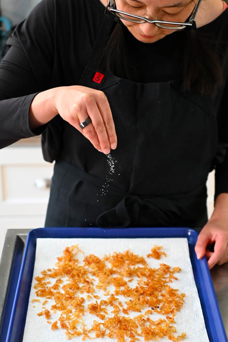 An Asian woman in a black apron is sprinkling salt on a tray of crispy shallots.