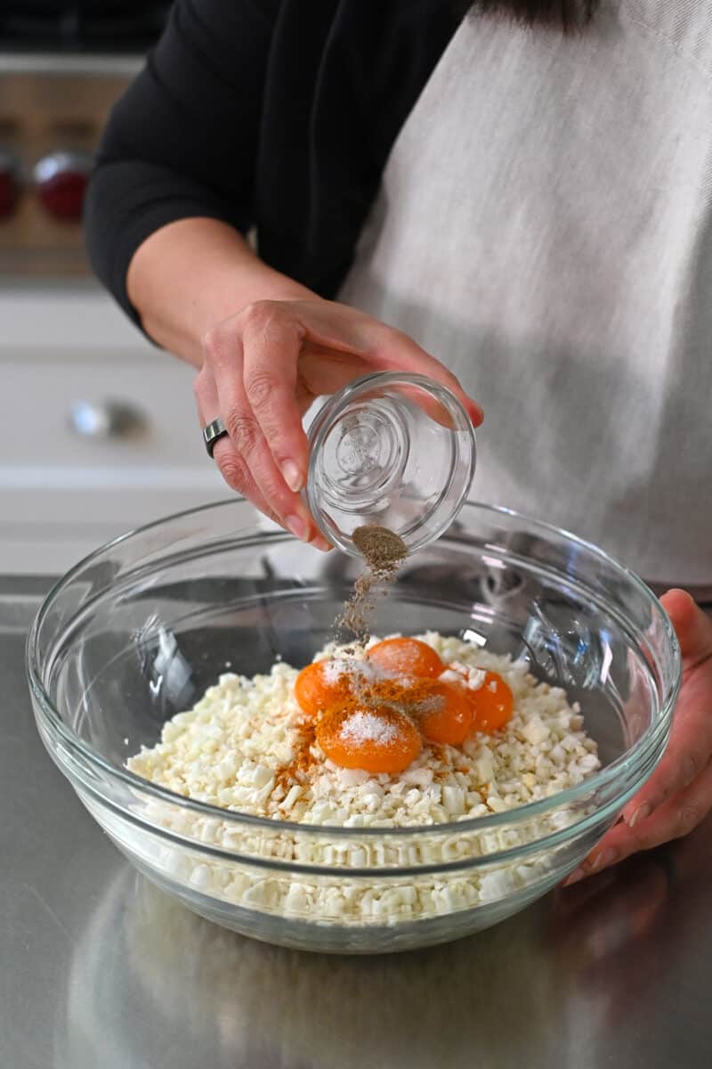 Ground white pepper is added to a large bowl filled with cauliflower rice, egg yolks, and salt.