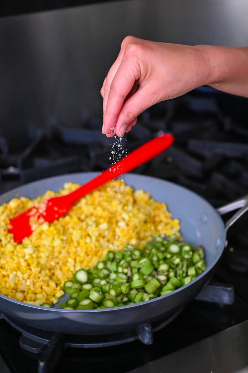 A hand is sprinkling salt on asparagus coins in a skillet filled with golden cauliflower fried rice.