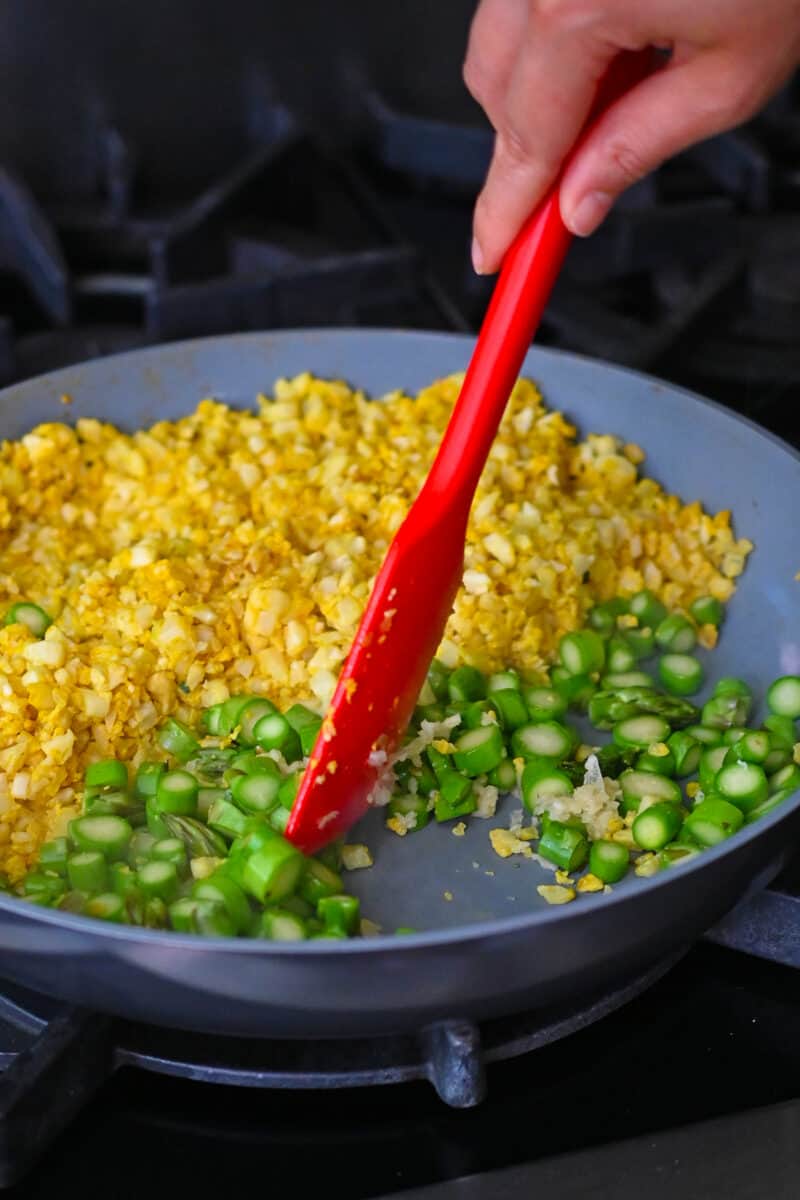 A red silicone spatula is mixing the asparagus coins with minced garlic in a pan that has yellow cauliflower rice in half of it.