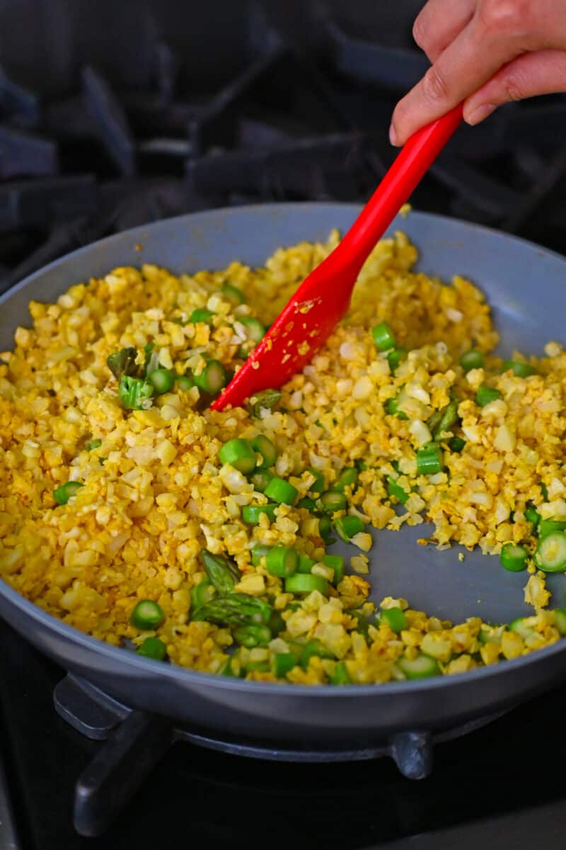 A red silicone spatula is stirring asparagus coins into the golden cauliflower fried rice.