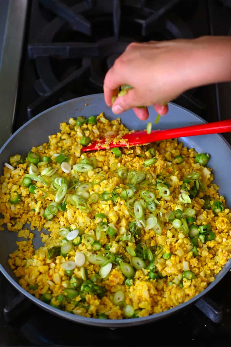 A hand is sprinkling sliced green onions onto a skillet filed with golden cauliflower fried rice.