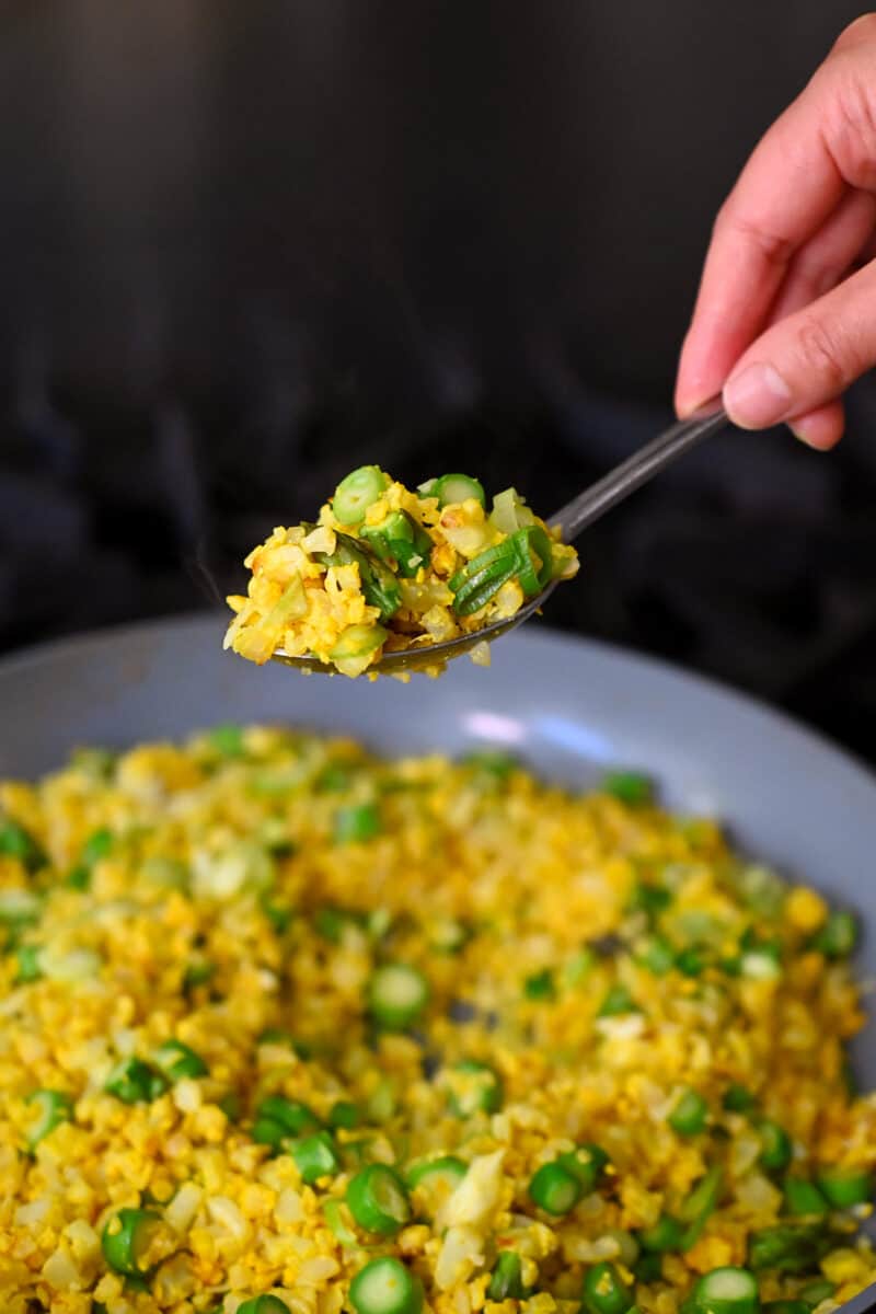 A spoon lifting up a scoop of golden cauliflower fried rice from a skillet.