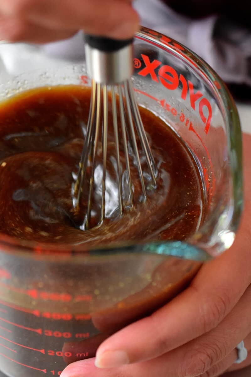 A closeup of a whisk stirring a brown liquid in a glass measuring cup.