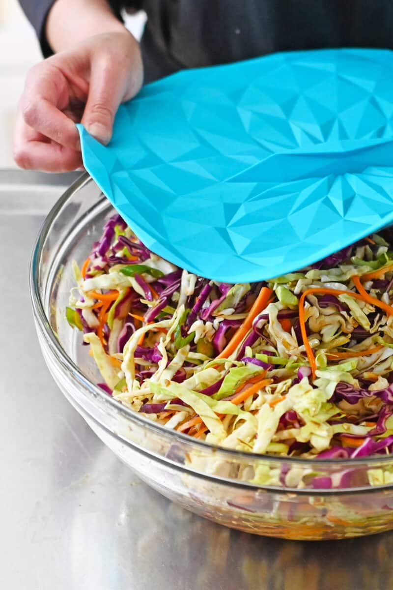 Placing a silicone lid on a glass bowl filled with pineapple coleslaw.