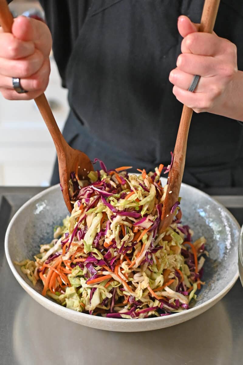 Tossing pineapple coleslaw with wooden spoons in a ceramic salad serving bowl. 