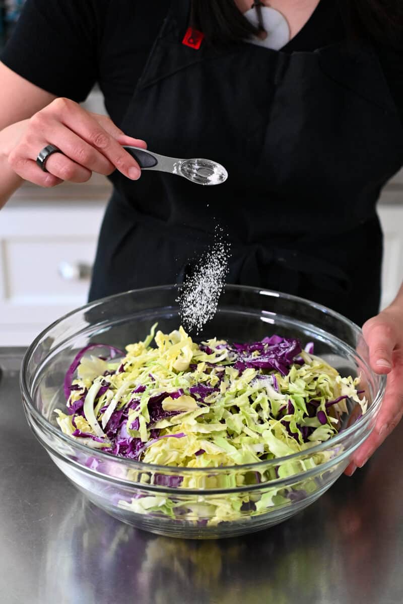Adding salt to a bowl of shredded green and purple cabbage.