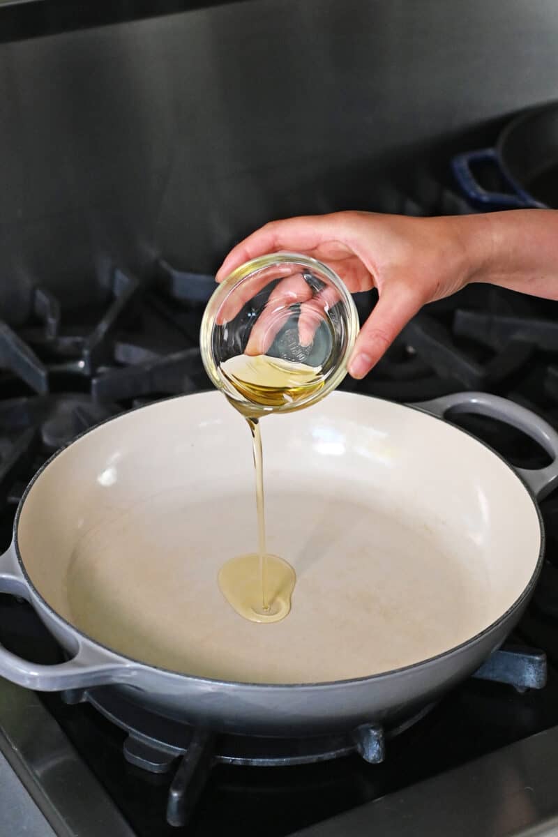 A hand is pouring a small bowl of avocado oil into a large enamel cast iron skillet.