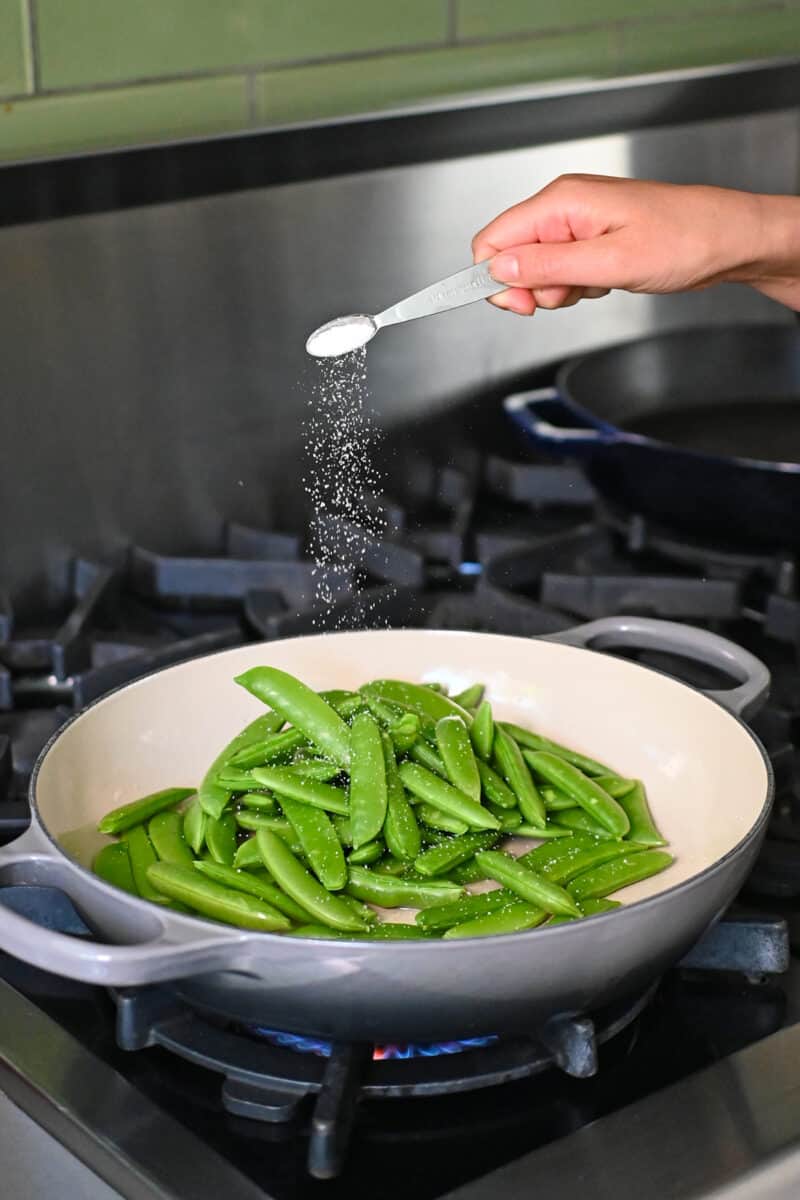 Adding a small spoonful of salt into a skillet with sugar snap peas.