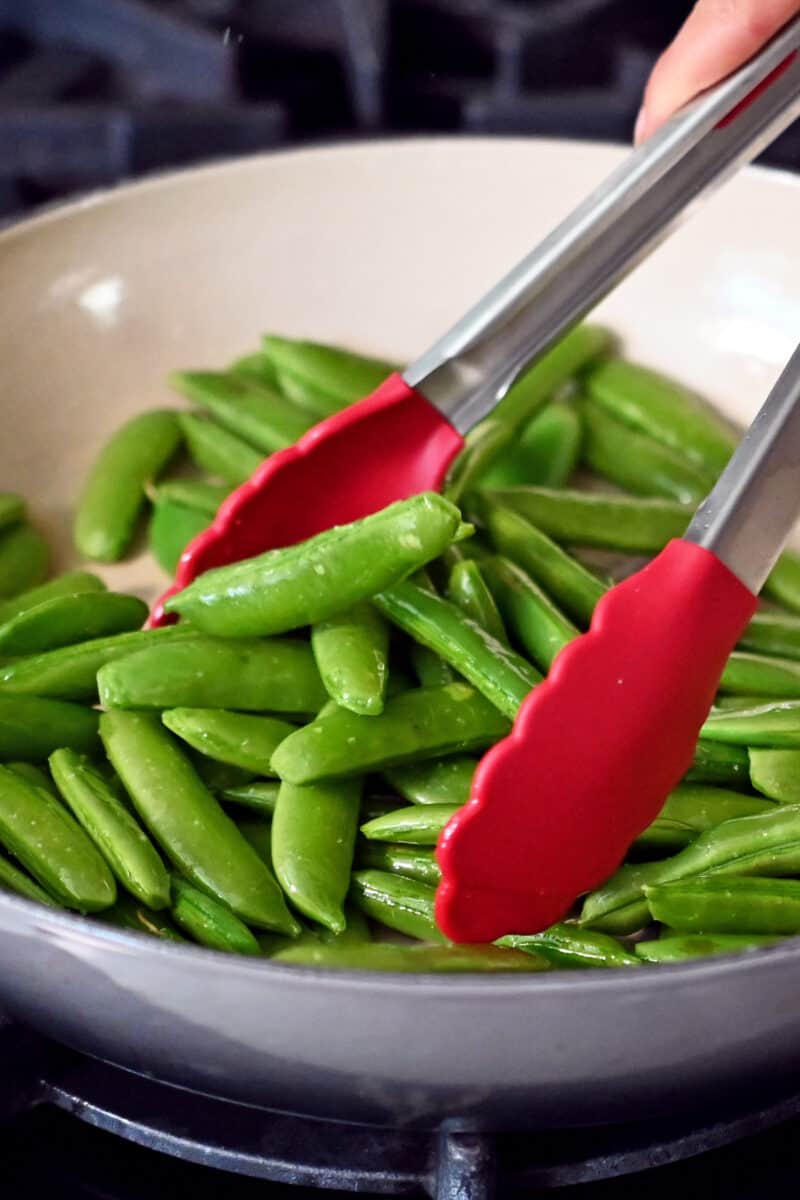 Tossing bright green sugar snap peas in a large skillet with a pair of tongs.