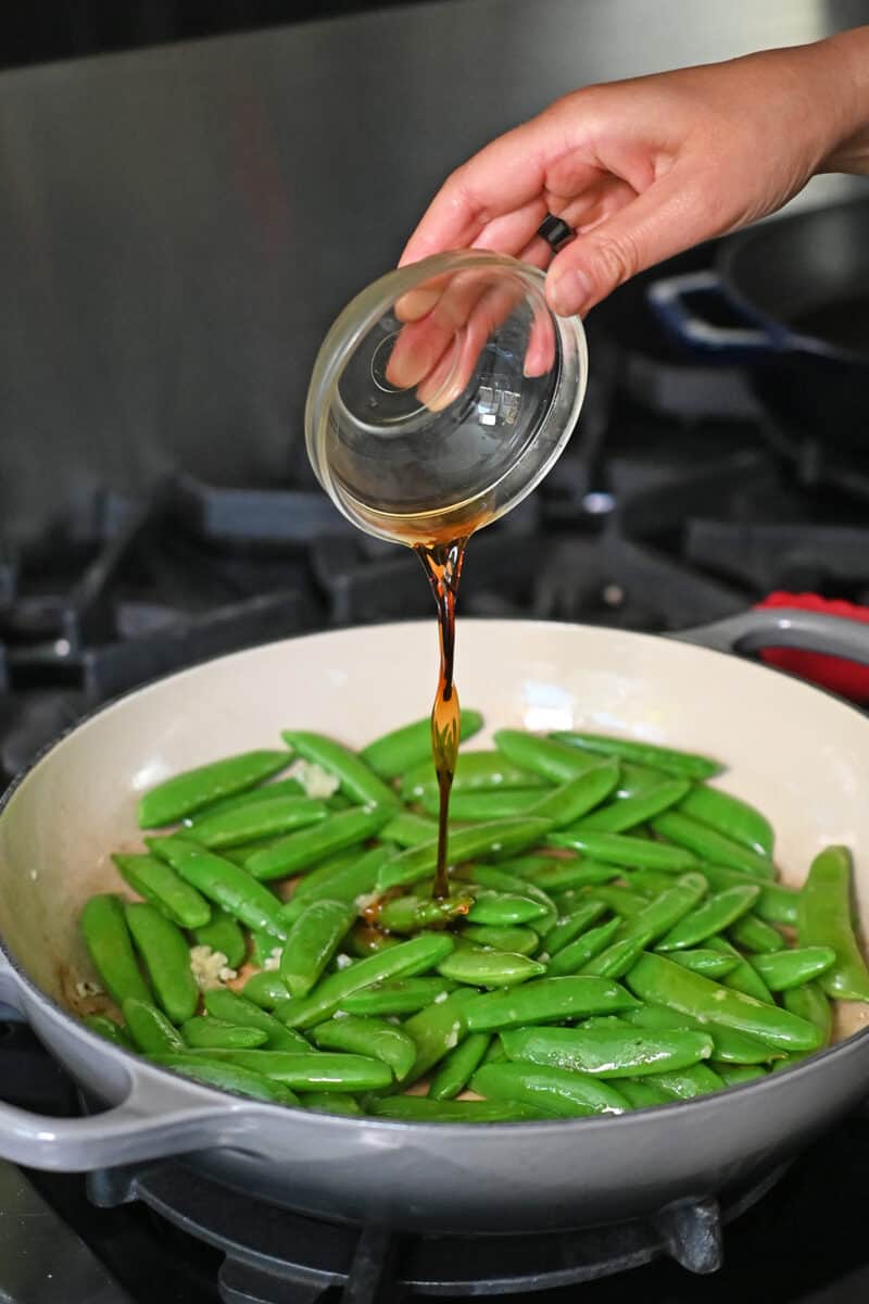 Pouring a small bowl of fish sauce into a skillet filled with sugar snap peas and garlic.