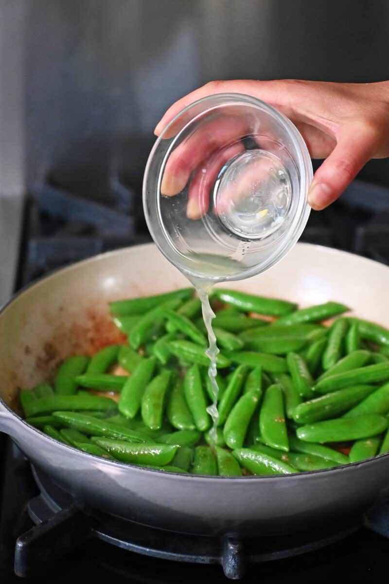Pouring a small bowl of freshly squeezed lime juice into a skillet with sugar snap peas, garlic, and fish sauce.