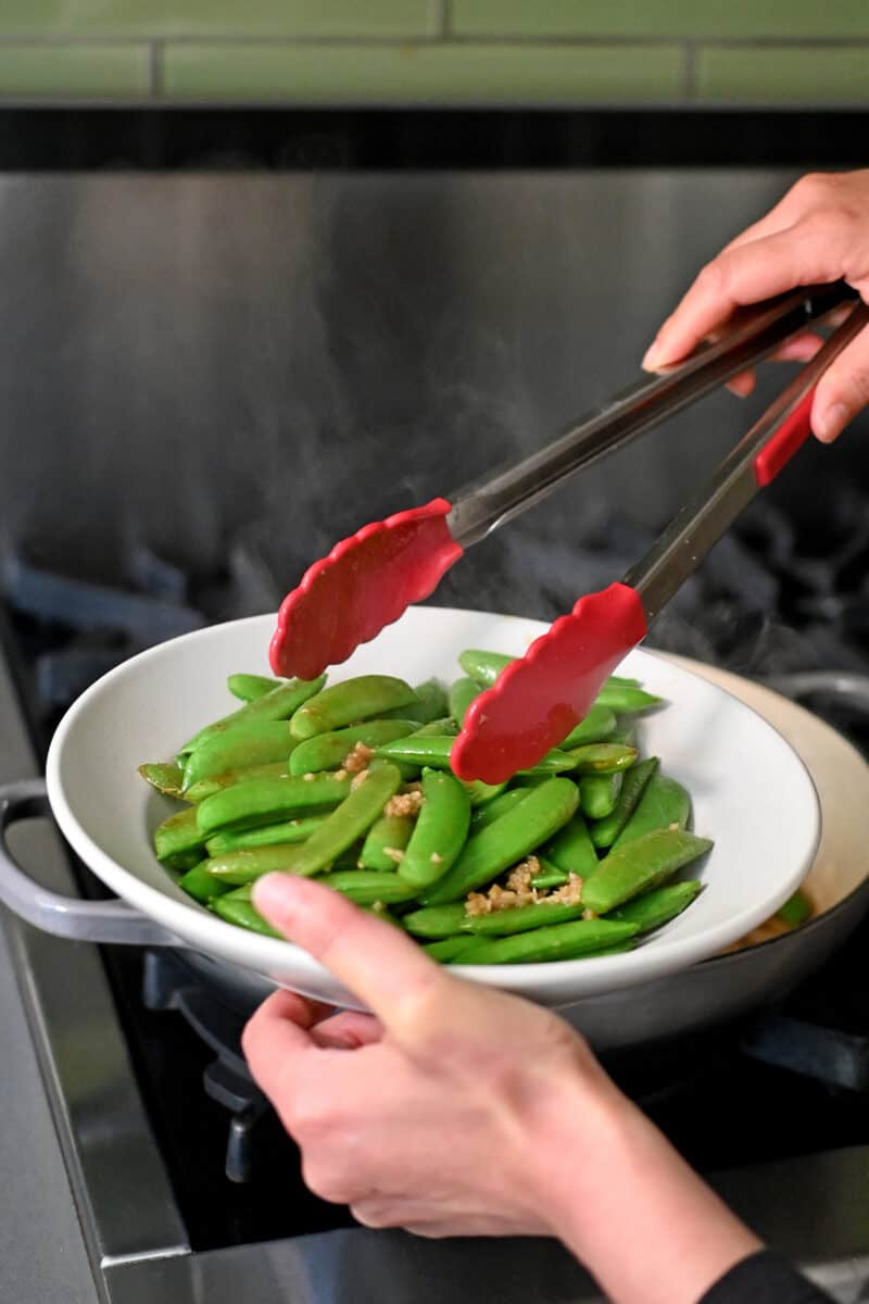 Using a pair of tongs to transfer sautéed sugar snap peas to a shallow off-white bowl.