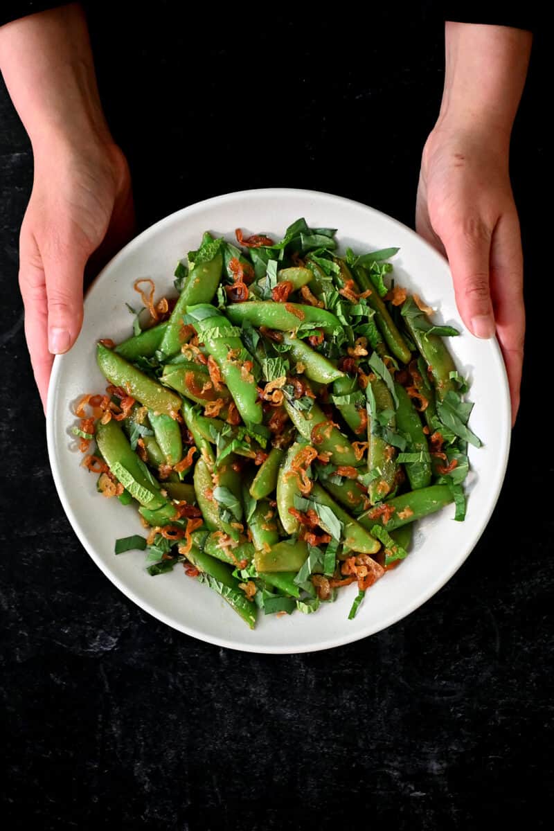 Two hands are holding a shallow dish filled with sautéed sugar snap peas with mint, basil, lime juice, and crispy shallots.