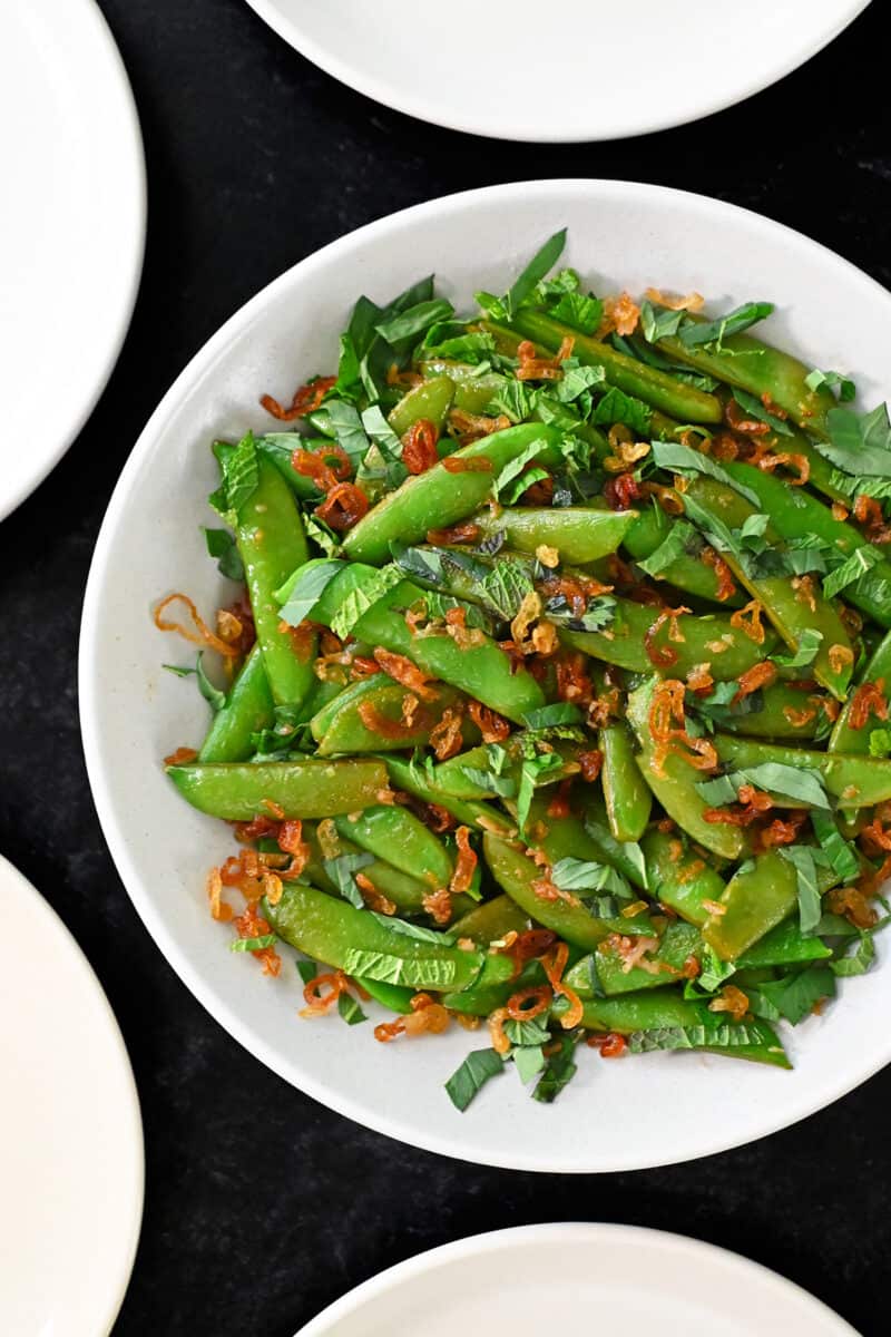 An overhead shot of a bowl of Asian sugar snap peas topped with fresh herbs and crispy shallots.