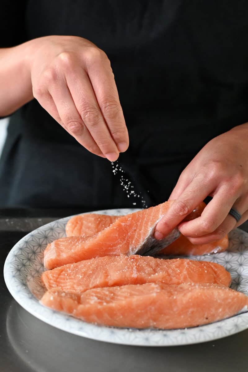 Sprinkling salt on four raw salmon fillets on a white plate.
