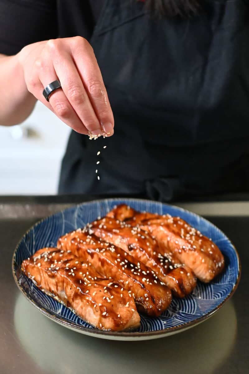 Sprinkling toasted sesame seeds on a plate with air fryer salmon fillets with honey sriracha fillets.