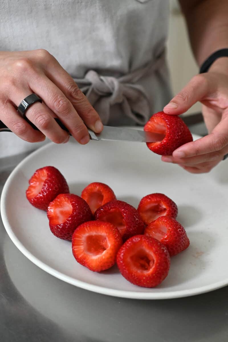 A paring knife is hollowing our a strawberry on top of a plate filled with hollowed out strawberries.