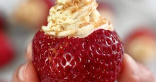 A hand is holding up a paleo and vegan cheesecake stuffed strawberry topped with gluten-free cookie crumbs.