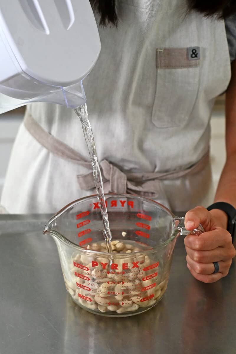 Pouring water into a glass measuring cup filled with raw cashews.