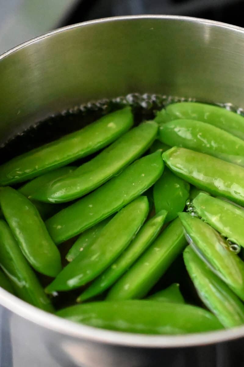 An small saucepan is filled with boiling water and bright green sugar snap peas.
