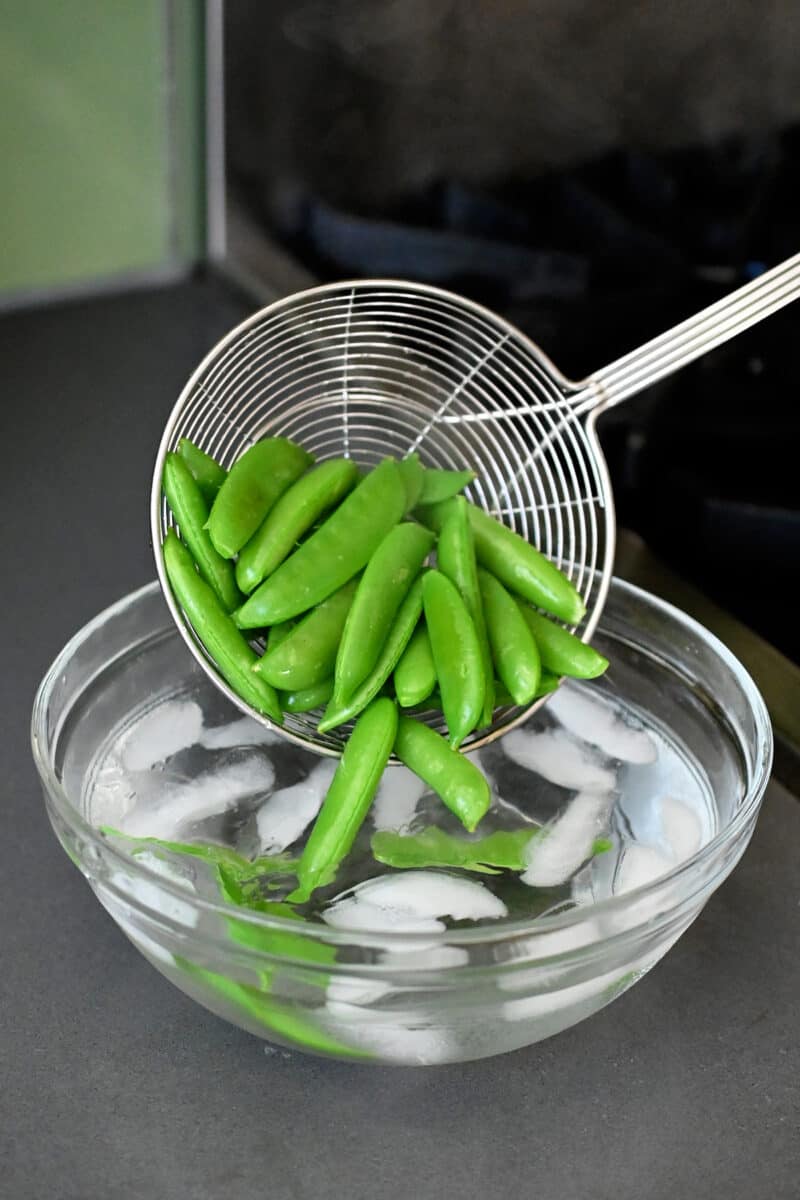 A metal strainer is adding cooked sugar snap peas to a bowl of ice water.
