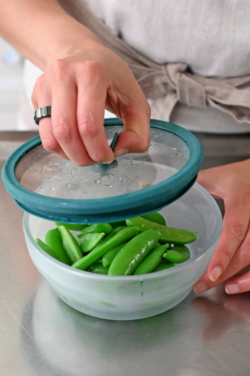 A hand is opening a glass microwave container filed with bright green, cooked sugar snap peas.