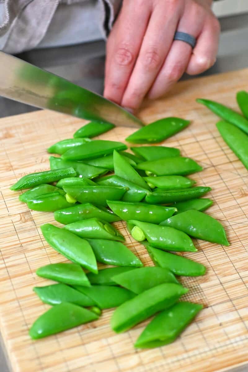 Chopping blanched sugar snap peas in half on a wooden cutting board.