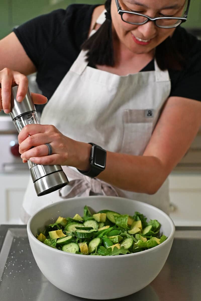 An Asian woman is adding freshly cracked black pepper to a large bowl of chopped salad.
