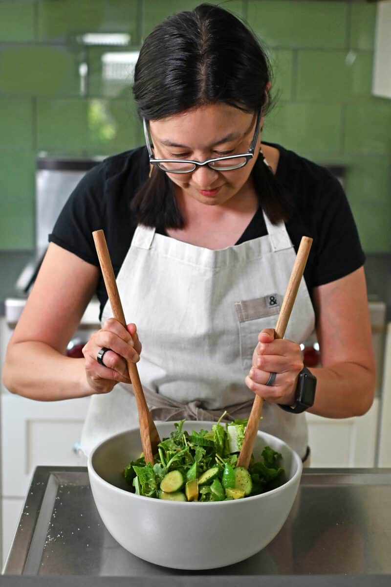 An Asian woman tossing a chopped salad with sesame ginger dressing in a large white bowl.