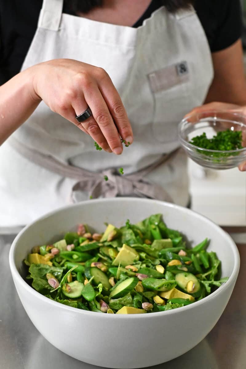 A hand is sprinkling sliced chives on top of a large bowl filled with chopped salad and sesame ginger dressing.