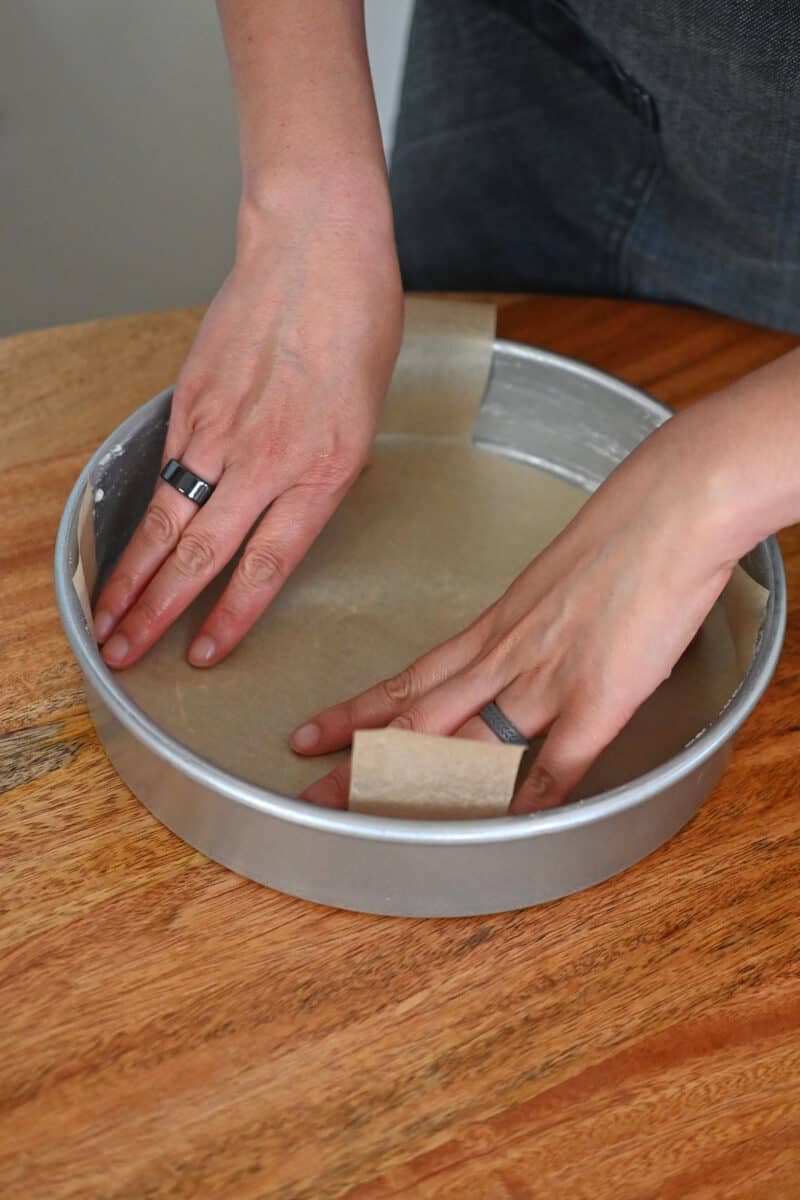Two hands are pressing a piece of parchment paper into a greased round cake pan.