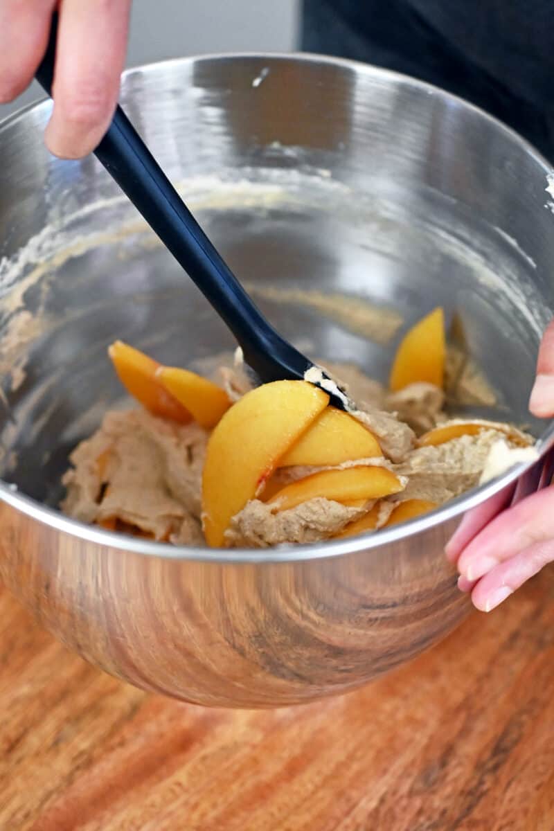 Folding peach slices into the cake batter with a black silicone spatula.
