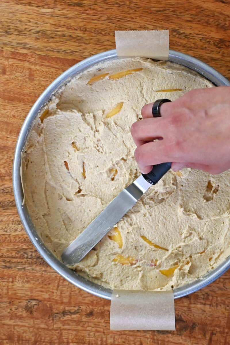 An overhead shot of someone using an offset spatula to smooth the top of an unbaked paleo peach cake.