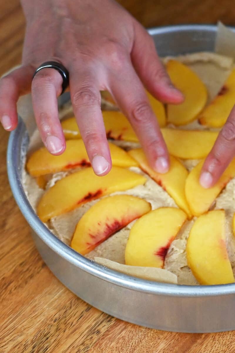 A hand is arranging peach slices on top of an unbaked cake.