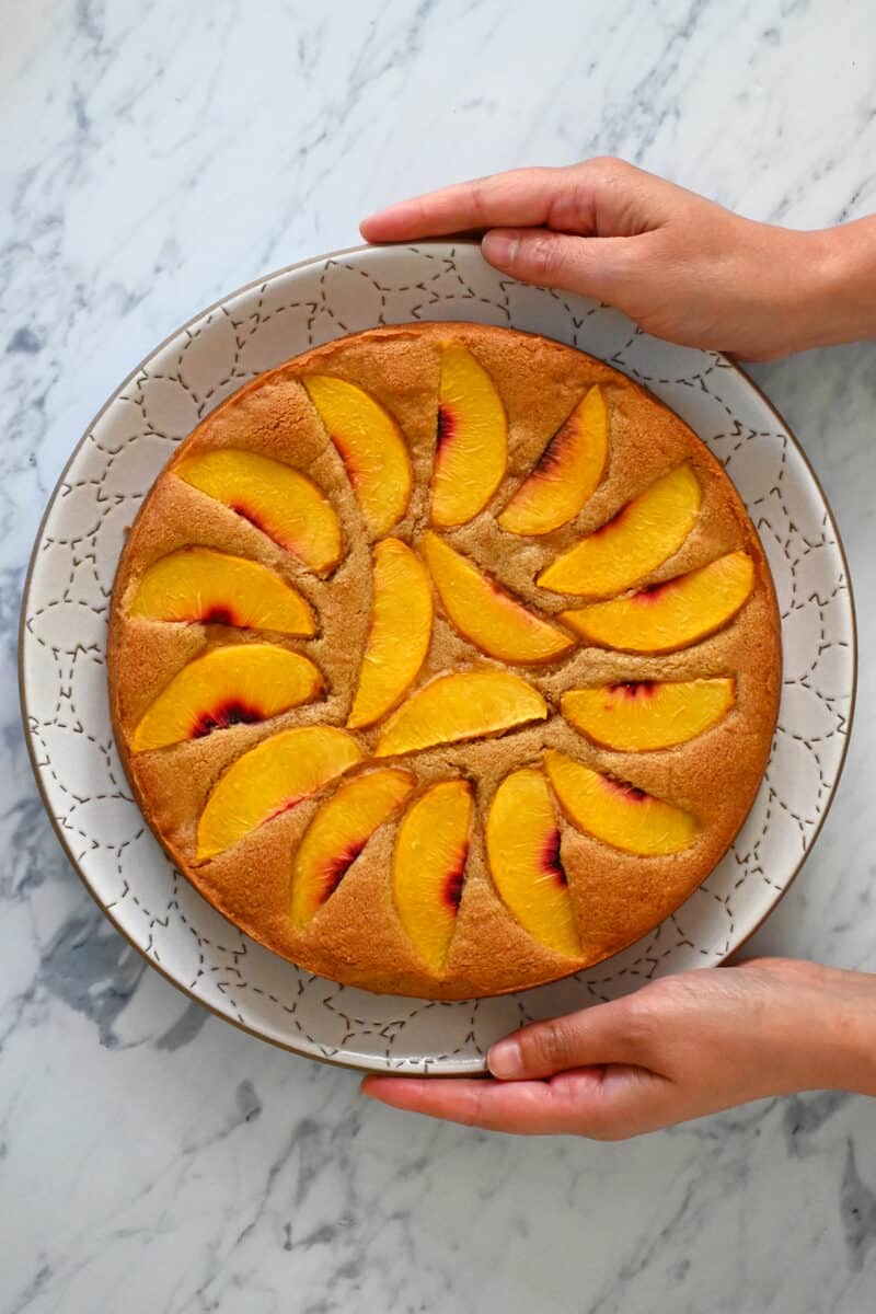 An overhead shot of a paleo peach cake on a white plate being held by two hands.