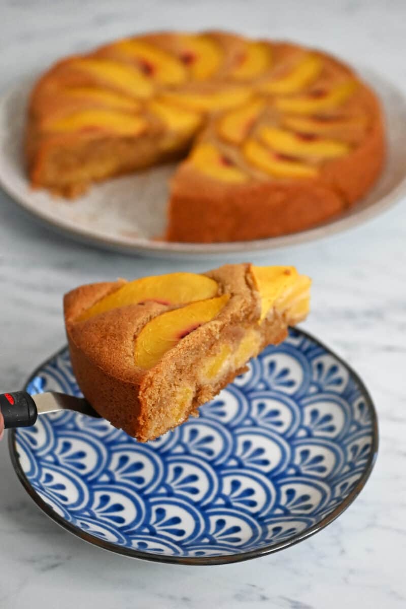 A slice of gluten free and paleo peach cake is being placed on a blue and wihite dish.