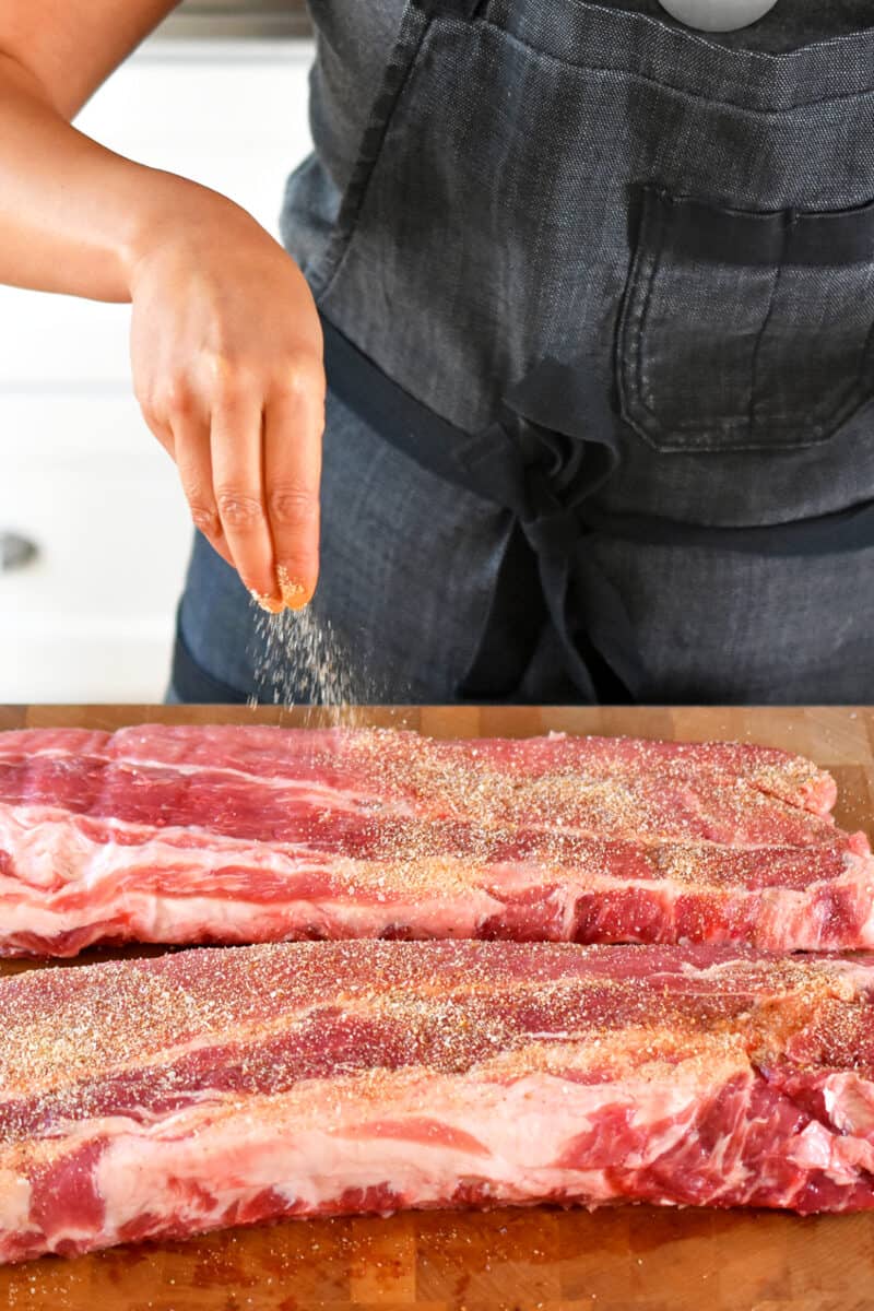 A person in a gray apron is sprinkling sugar-free dry rub on baby back ribs.