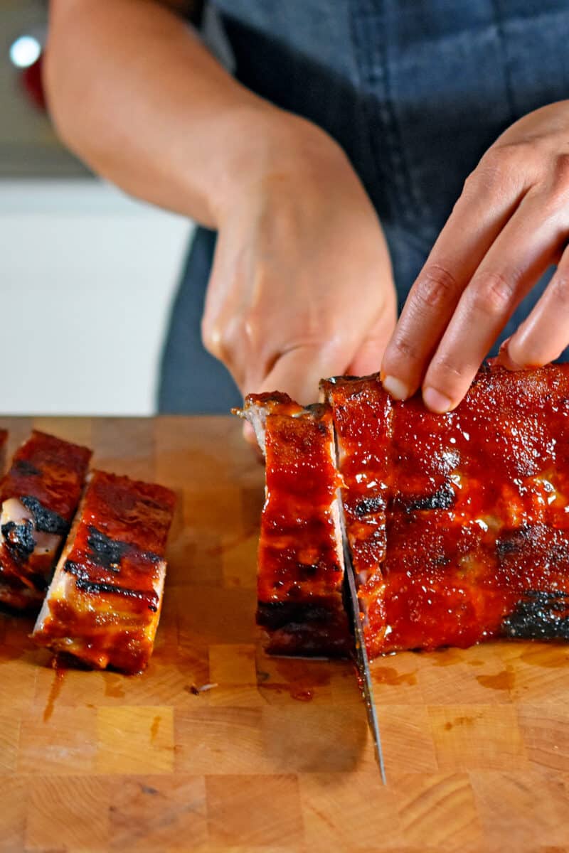 Slicing oven baked baby back ribs with a chef's knife on a wooden cutting board.