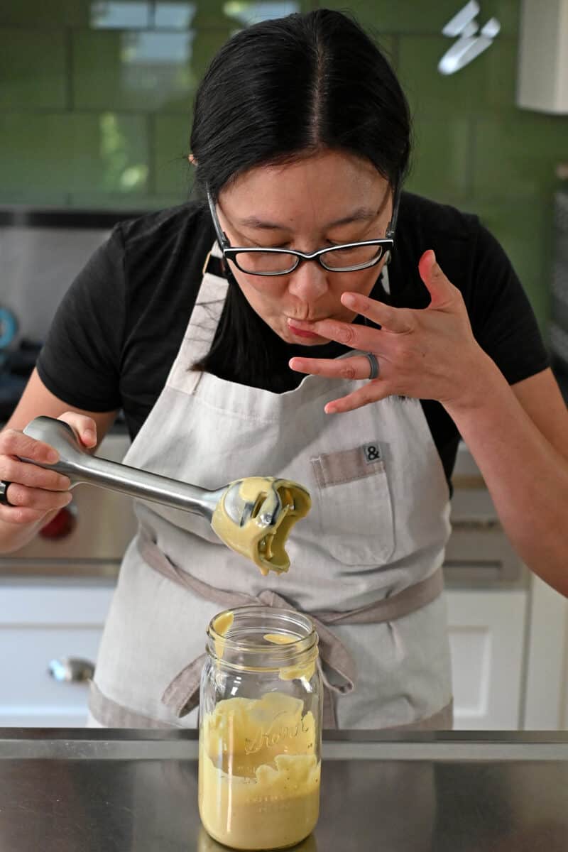 An Asian woman with glasses is tasting a freshly blended batch of dairy free caesar dressing.