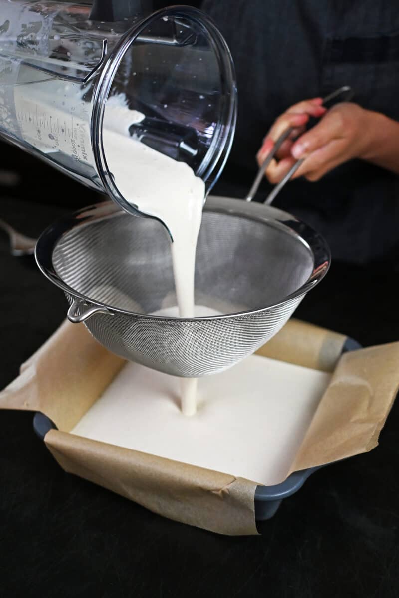 Pouring the coconut cream bar filling through a fine mesh sieve onto the crust in a square pan.