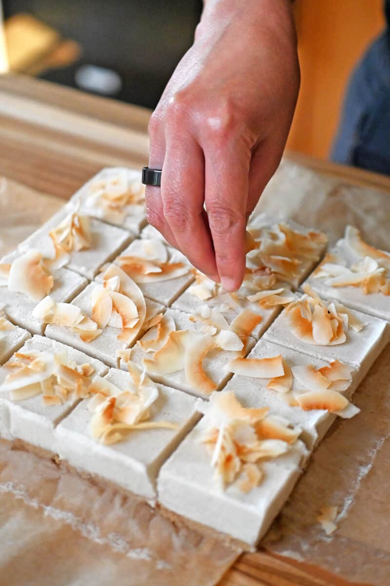 A hand adding toasted coconut flakes to squares of coconut cream bars.