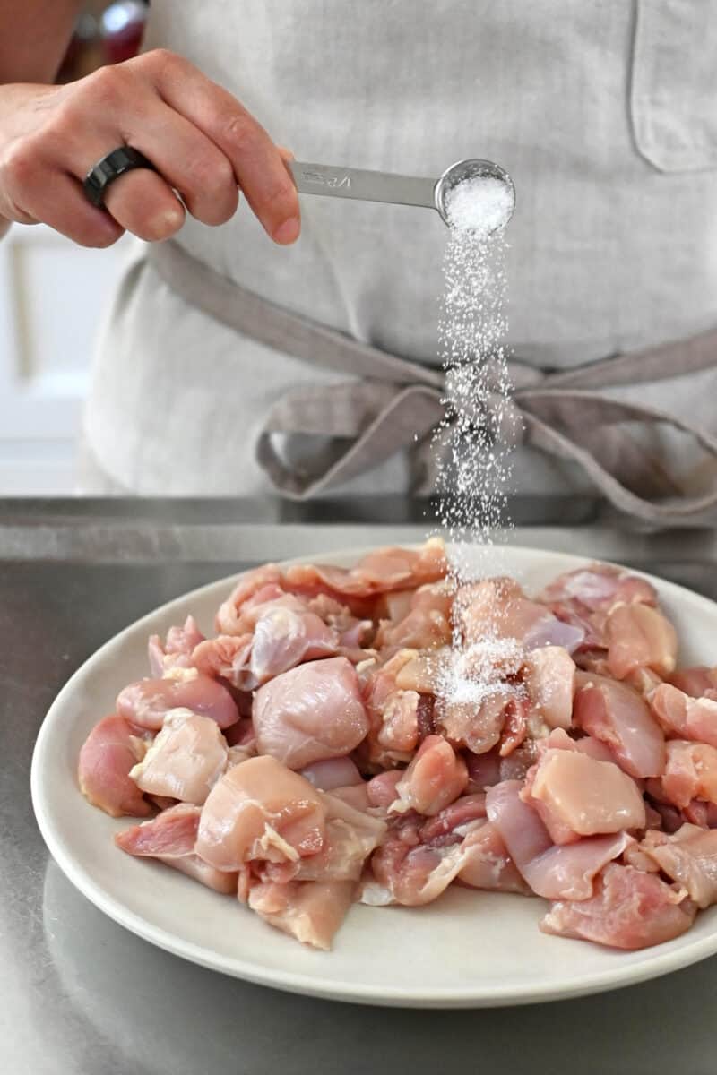 A person in an off-white apron is adding a small spoonful of salt to a plate filled with raw cut-up boneless, skinless chicken thighs.