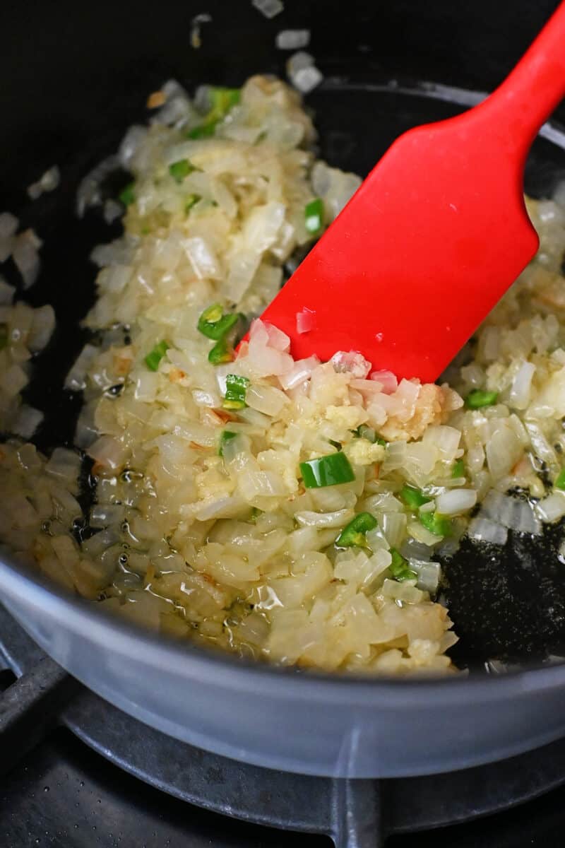 A red silicone spatula is mixing sautéed chopped onions, minced ginger, minced garlic, and diced green peppers in a pot.