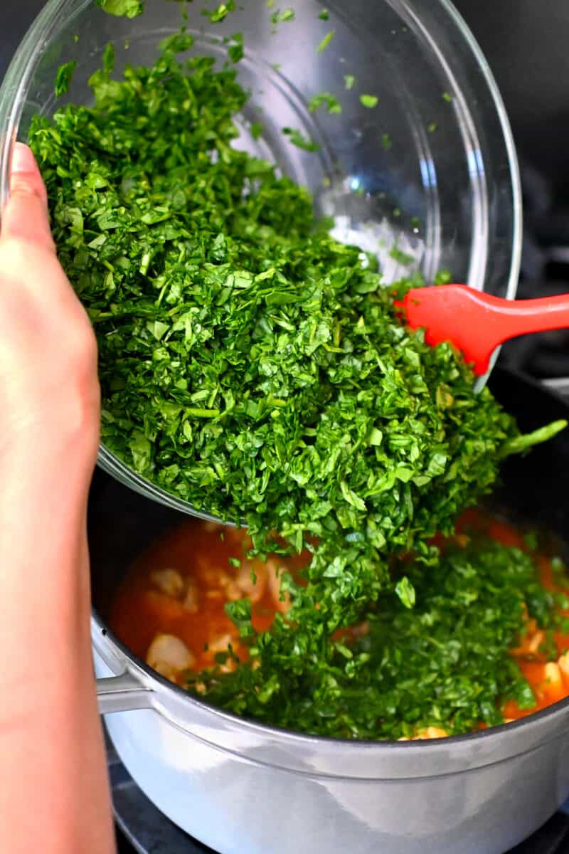 Adding a big bowl of chopped baby spinach to a pot filled with chicken and tomato curry.