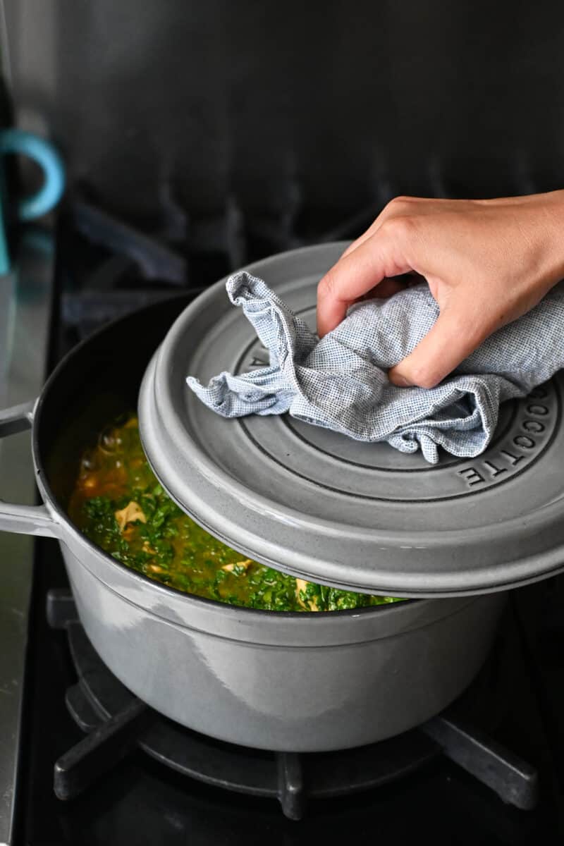 A hand clasping a cloth is putting the lid on a gray dutch oven filled with chicken saag.