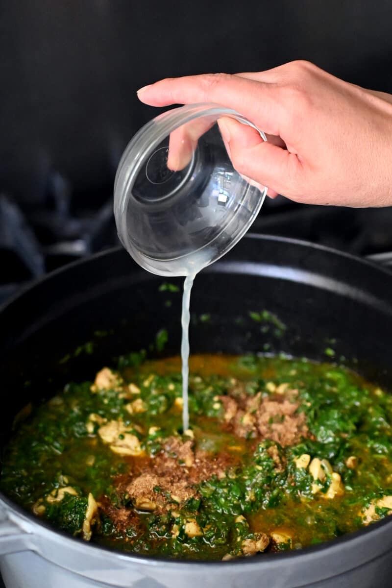 Pouring a small bowl of lemon juice into a pot filled with cooked chicken saag curry.