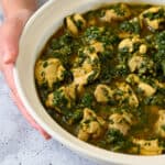 A hand is holding an off-white bowl filled with chicken and spinach Indian curry.