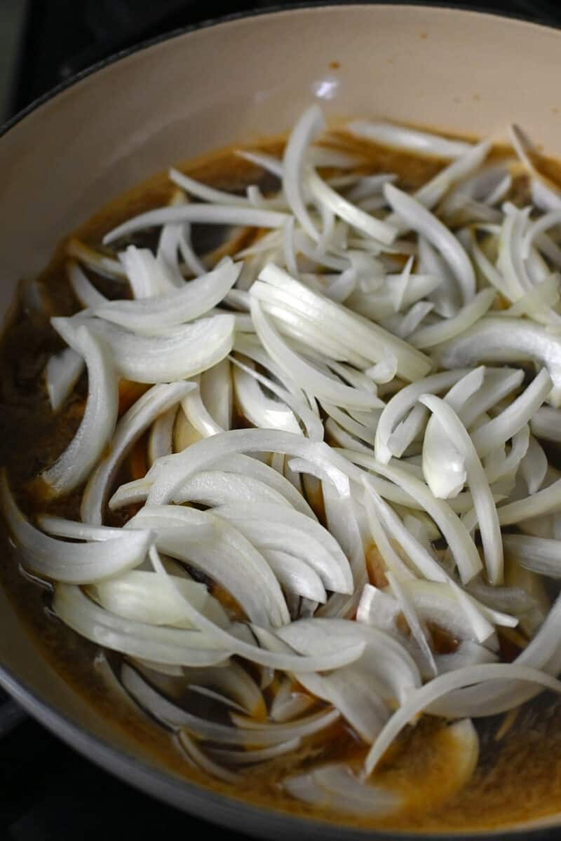 A closeup shot of raw thinly sliced onions in an enameled cast iron skillet in a brown sauce.
