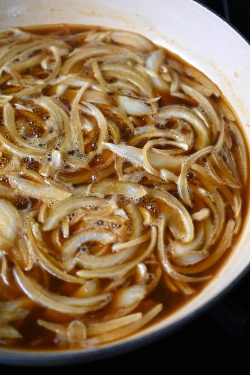 A large enamel cast iron skillet filled with thinly sliced onions in a simmering brown sauce. 